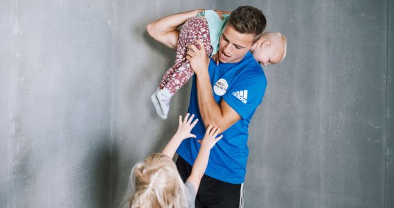 Ein kleines Mädchen streckt sich zu einem junger Mann in Sportkleidung, der ein lachendes Kleinkind über der Schulter hält