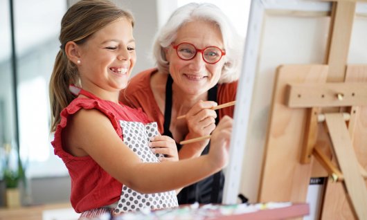 Ein kleines, lachendes Mädchen und eine alte Frau malen gemeinsam ein Bild auf die Leinwand