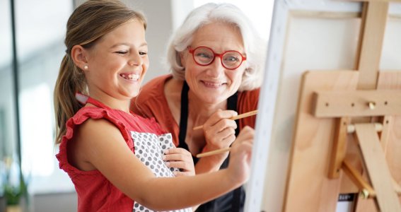 Ein kleines, lachendes Mädchen und eine alte Frau malen gemeinsam ein Bild auf die Leinwand