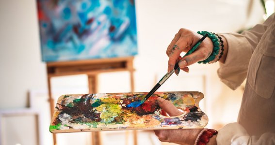 Eine junge Künstlerin bereitet Malfarben auf einer Palette vor, im Hintergrund steht eine Leinwand mit abstrakter Malerei