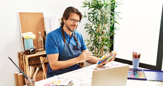 Ein lächelnder Mann hält einen Pinsel und eine Farbpalette und schaut auf einen Laptop