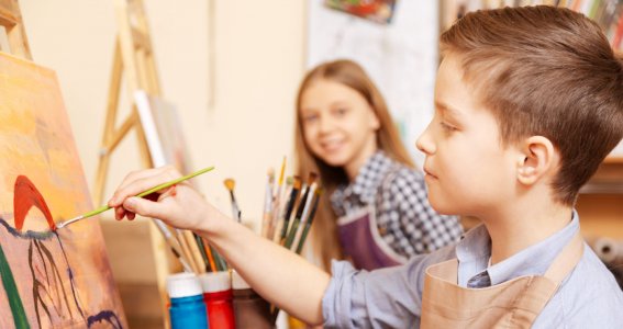 Ein Junge malt konzentriert vor einer Staffelei und ein Mädchen im Hintergrund lächelt