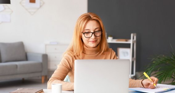 Eine Frau sitzt im Wohnzimmer vor dem Laptop und schreibt Notizen