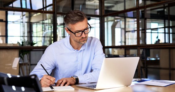 Ein Mann sitzt in einem Raum mit Glaswänden und schreibt vor dem Laptop Notizen