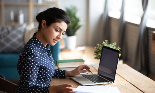 Excel lernen für Fortgeschrittene Online Eine junge Frau sitzt an einem Holztisch vor einem Laptop und blättert in einem Notizblock