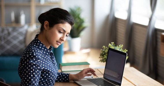 Eine junge Frau sitzt an einem Holztisch vor einem Laptop und blättert in einem Notizblock