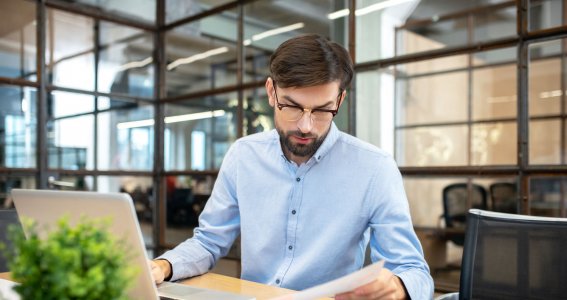 Ein Mann sitzt in einem Raum mit Glaswänden vor einem Laptop und schaut auf ein Blatt Papier