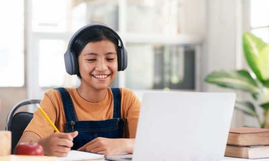 Computerkurs für Kinder Online Ein lachendes Mädchen sitzt mit Kopfhörern vor dem Laptop und macht sich Notizen