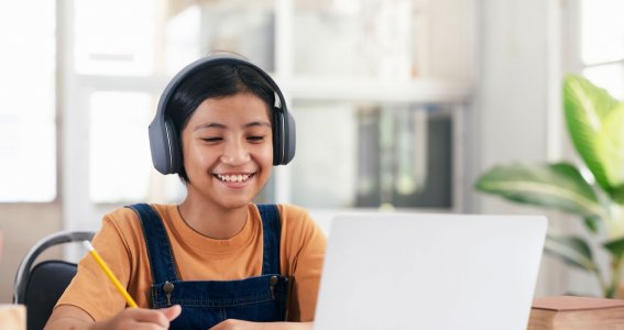 Ein lachendes Mädchen sitzt mit Kopfhörern vor dem Laptop und macht sich Notizen