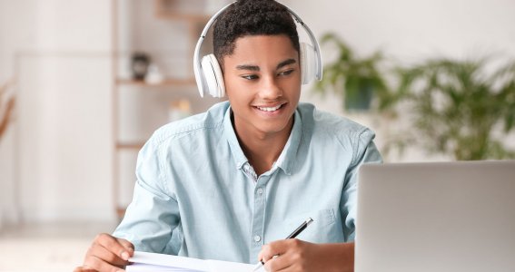 Ein lächelnder Teenager sitzt mit weißen Kopfhörern vor dem Laptop und schreibt auf Papier