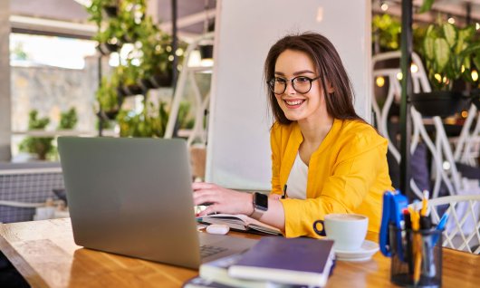 EDV Kenntnisse lernen für das Studium Eine lachende junge Frau sitzt am Holztisch vor dem Laptop und einigen Büchern