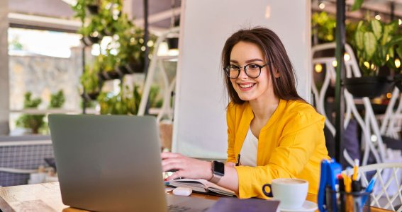 Eine lachende junge Frau sitzt am Holztisch vor dem Laptop und einigen Büchern