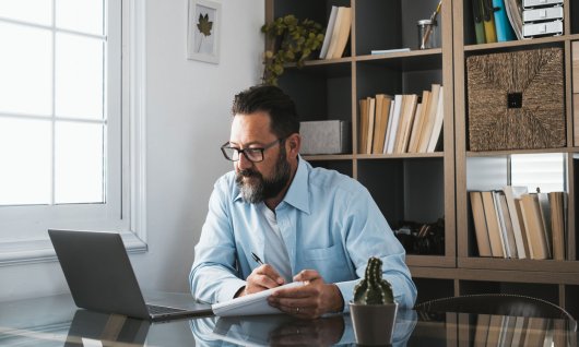 Excel lernen Online für Erwachsene Ein Mann mittleren Alters sitzt am Tisch vor einem Laptop und macht sich Notizen
