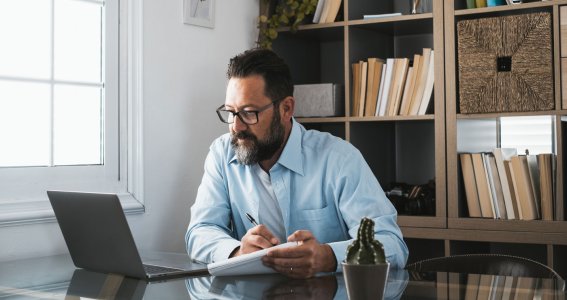 Ein Mann mittleren Alters sitzt am Tisch vor einem Laptop und macht sich Notizen