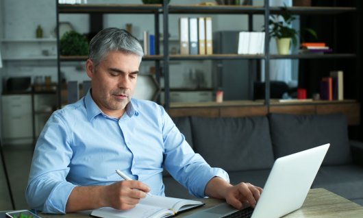 Ein Mann mit grauen Haaren sitzt im Wohnzimmer vor dem Laptop und schreibt in sein Notizbuch