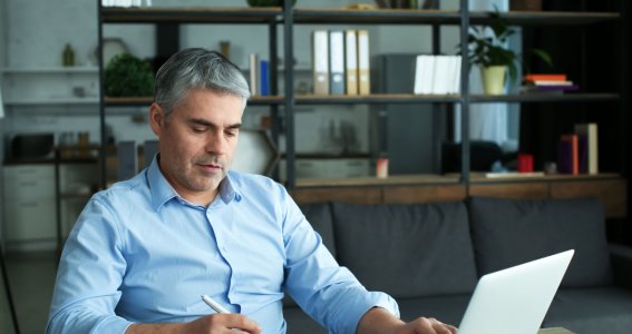 Ein Mann mit grauen Haaren sitzt im Wohnzimmer vor dem Laptop und schreibt in sein Notizbuch