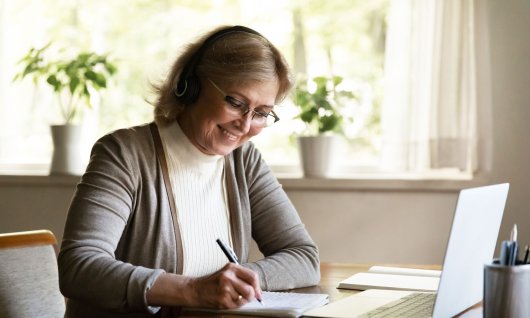 Computerkurs für Senioren online Eine ältere Frau sitzt lachend im Wohnzimmer vor einem Laptop und schreibt auf Papier