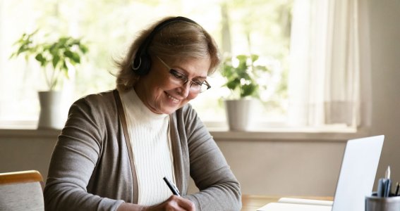 Eine ältere Frau sitzt lachend im Wohnzimmer vor einem Laptop und schreibt auf Papier