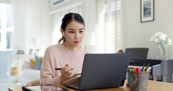 Eine junge Frau schreibt in den Notizblock und sitzt vor dem Laptop im Wohnzimmer