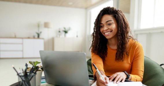 Eine junge, lachende Frau mit lockigen Haaren sitzt im Sofasessel und schreibt Notizen vor dem Laptop