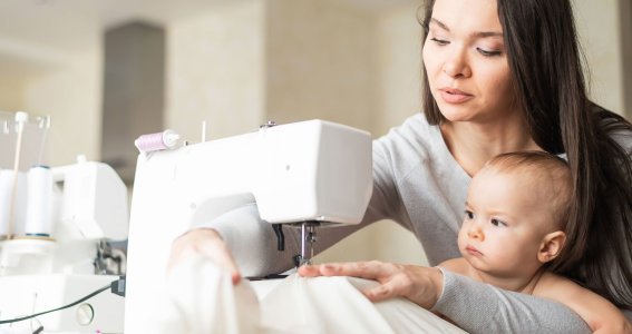 Frau sitzt mit Baby vor der Nähmaschine. 