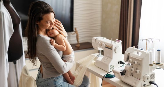Frau sitzt mit Baby vor einer Nähmaschine. 
