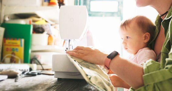 Frau sitzt mit Baby vor einer Nähmaschine. 