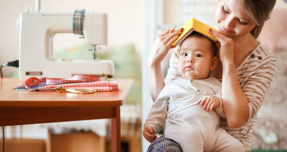 Frau sitz mit Baby und Buch auf dem Kopf vor einer Nähmaschine. 