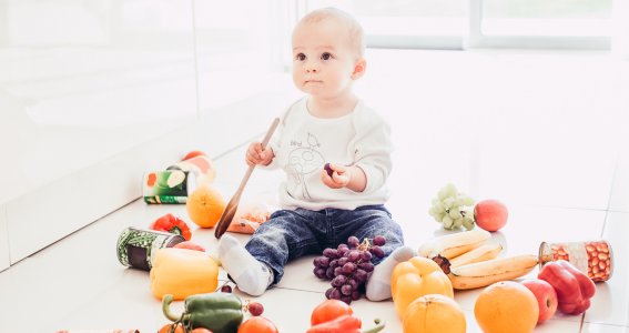 Baby mit Kochlöffel sitzt auf den Küchenboden umgeben von Obst und Gemüse