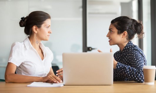 Zwei Frauen sitzen nebeneinander an einem Tisch mit Laptop und unterhalten sich