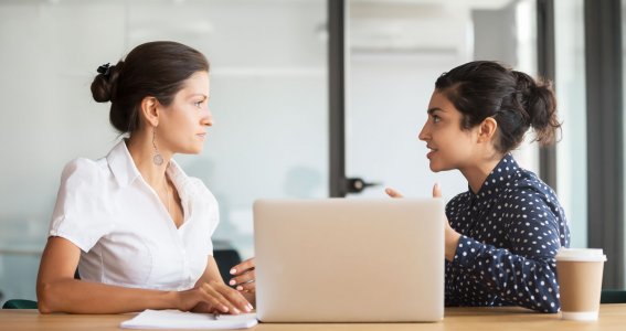 Zwei Frauen sitzen nebeneinander an einem Tisch mit Laptop und unterhalten sich