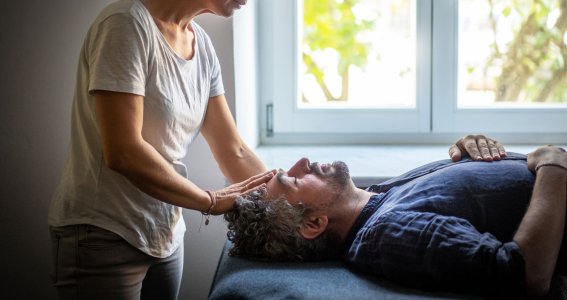 Eine Frau legt ihre Hand auf die Stirn eines Mannes, der auf einem Massagetisch liegt