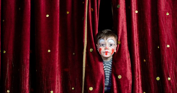 Junge steht mit Clownschminke zwischen Theatervorhängen. 