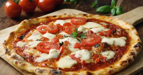 frische Pizza auf einem Holzblech mit Tomaten und Basilikum im Hintergrund