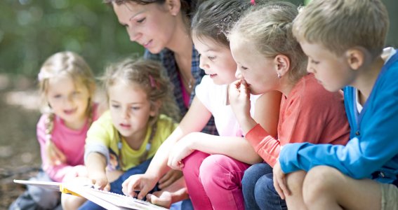 Kinder sitzen neben einer Frau und schauen mit ihr in ein Buch. 