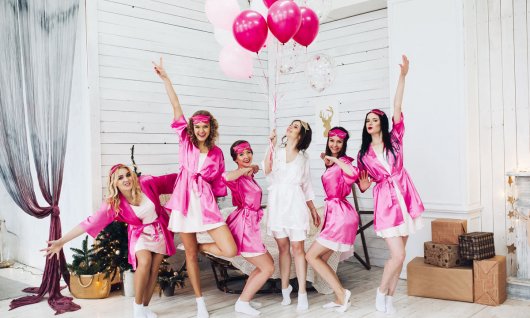 Eine Gruppe junger Frauen posieren feierlich in weißen Kleidern und pinken Bademänteln mit weiß-rosafarbenen Luftballons