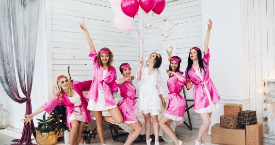 Eine Gruppe junger Frauen posieren feierlich in weißen Kleidern und pinken Bademänteln mit weiß-rosafarbenen Luftballons