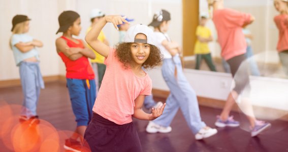 Ein Mädchen mit cooler Kappe in Hip Hop Pose und eine Gruppe von Kindern im Hintergrund im Tanzschritt
