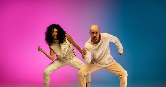 Ein Mann und eine Frau tanzen in legerer Kleidung vor einem blau-pinkem Hintergrund Hip Hop 