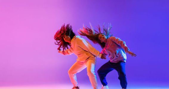Zwei junge Frauen mit wehenden langen Haaren tanzen vor einem blau-violetten Hintergrund Hip Hop Tanzschritte
