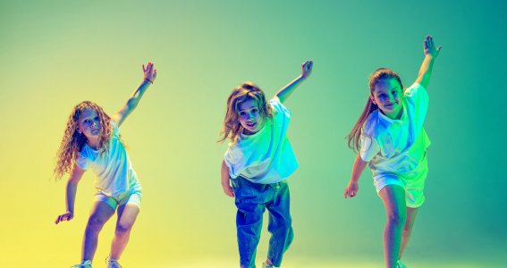 Drei tanzende Mädchen vor gelb-grünem Hintergrund beugen sich vor und strecken einen Arm hinter sich