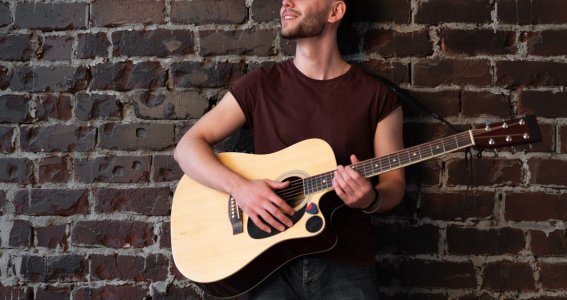 Ein junger Mann lächelt und lehnt an einer Ziegelwand mit einer Gitarre in der Hand