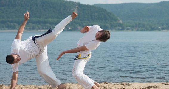 Zwei junge Männer in weißer Kleidung trainieren am Strand Capoeira