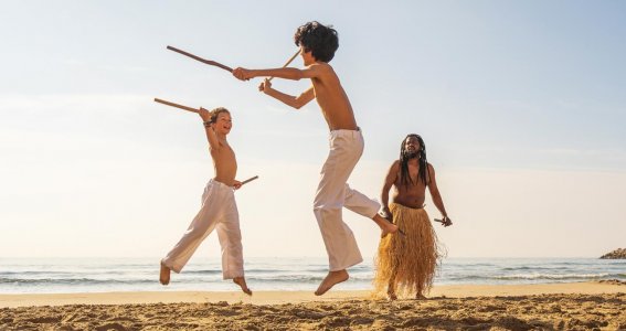Zwei Jungs in weißer Hose springen am Strand in die Höhe und halten Holzstöcke zum Angriff