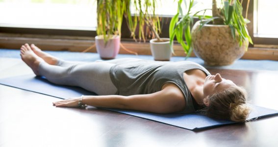 Eine Frau liegt auf einer Yogamatte neben Zimmerpflanzen und hat die Augen geschlossen