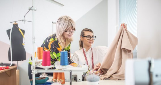 Zwei Frauen sind an einer Nähmaschine nähen und schauen Stoffe an. 