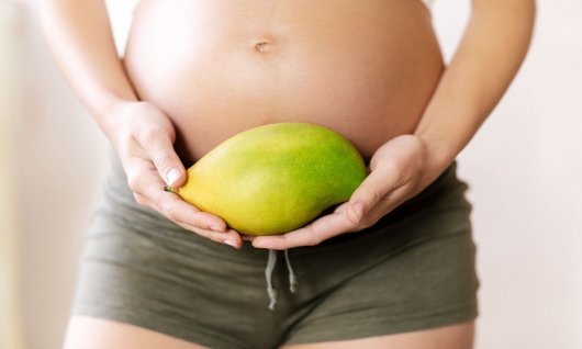 Schwangere Frau die eine Mango vor ihren Bauch hält.