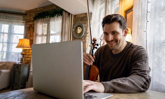 Mann halt eine Geige und sitzt vor seinem Laptop