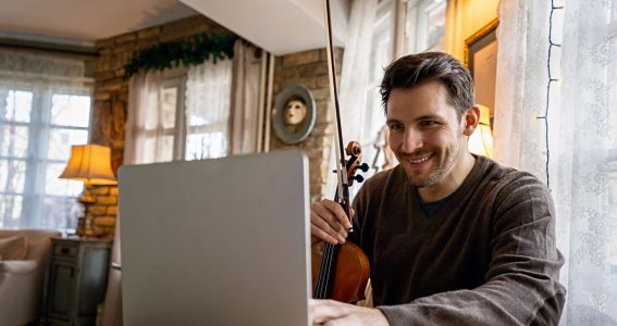 Mann halt eine Geige und sitzt vor seinem Laptop