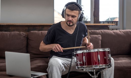 Online Percussionunterricht für Erwachsene Mann sitzt auf der Couch, spielt Trommel und schaut auf seinen Laptop